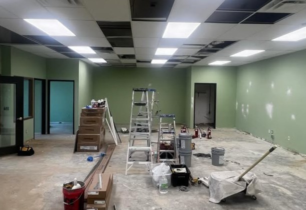 A room under renovation with a partially removed ceiling, green walls, and construction materials scattered. Ladders and paint cans suggest ongoing work.