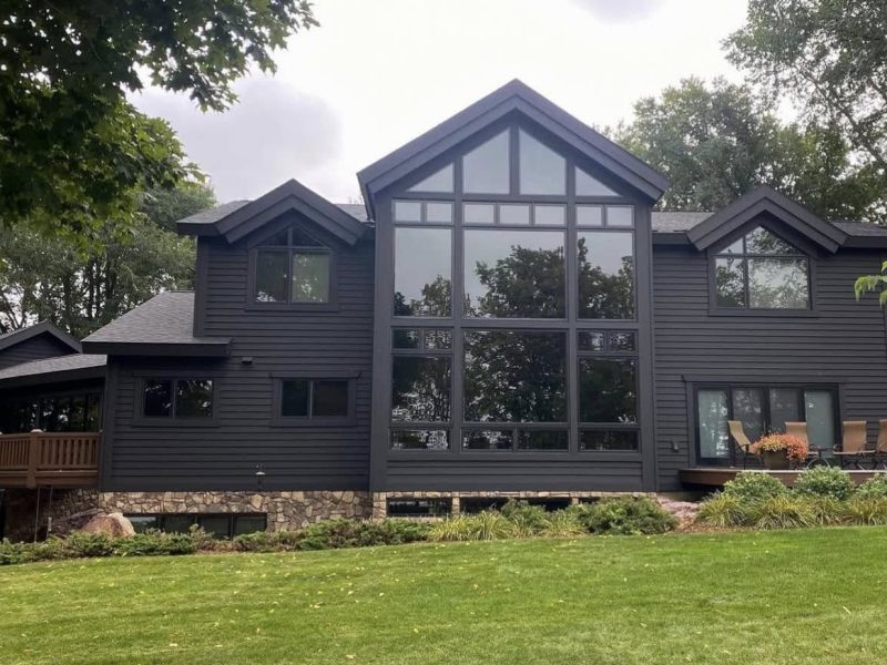 Modern dark gray house with large central windows reflecting trees. Surrounded by green lawn, patio with chairs, and lush foliage under cloudy sky.