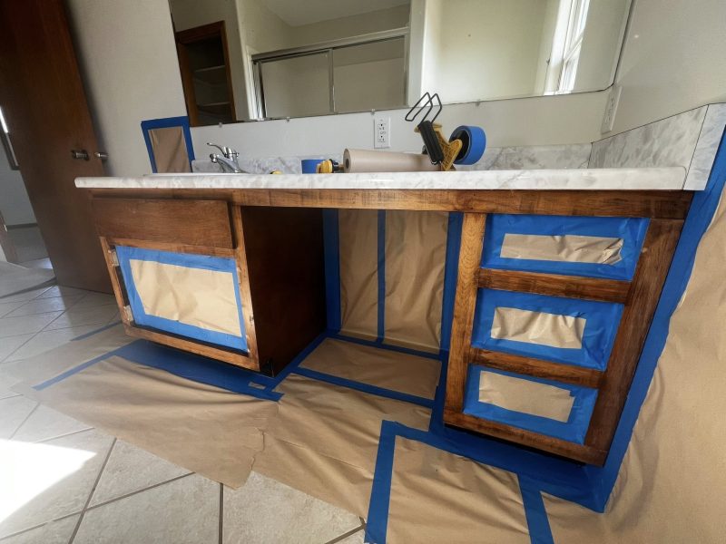 A wooden bathroom vanity is prepared for painting, with blue tape and brown paper covering its surfaces. Tools and tape rolls sit on a marble countertop.