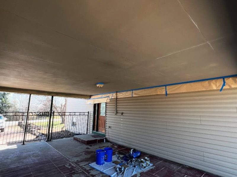 A covered patio under renovation features a taped ceiling with beige and blue masking materials. Painting supplies and buckets rest on a tarp below.