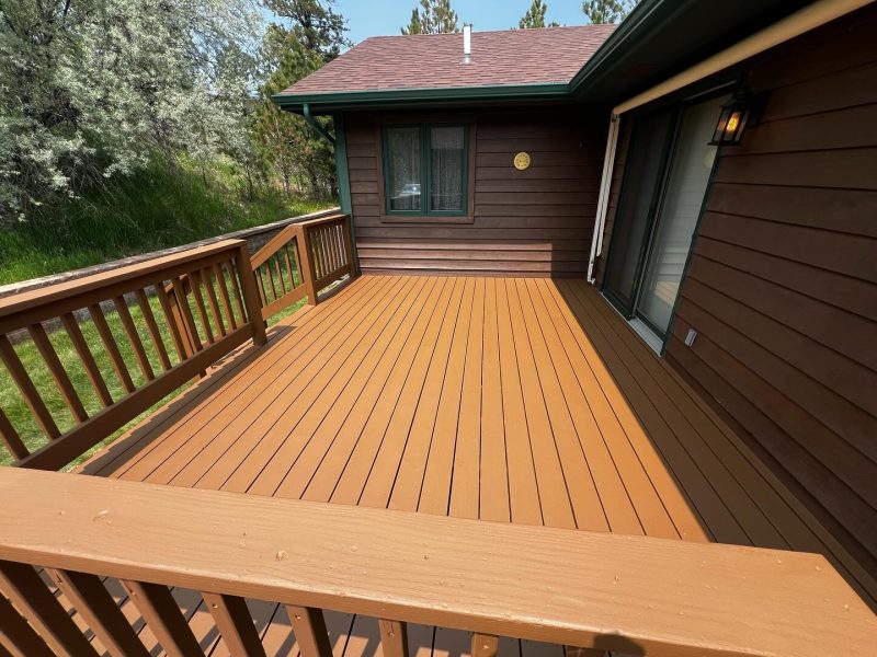 A wooden deck with a freshly stained, warm brown finish is attached to a house with dark wood siding. Green trees and grass are visible in the background.
