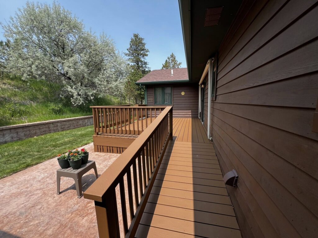 A wooden deck with railing extends from a brown house under clear blue skies. Potted flowers sit on a patio; lush green trees and grass surround the area.