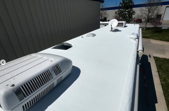 The image shows the white roof of an RV with air conditioning units and a satellite dish under a clear blue sky, next to a large building and trees.