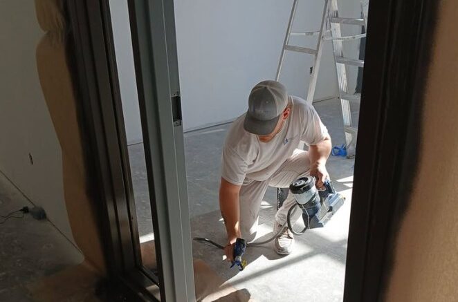 A person in a white outfit and cap uses a spray gun to paint a door frame. A tall ladder is visible in the background of the sunlit room.